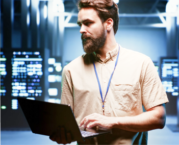 Person with lanyard using a laptop in a server room, with glowing data screens and equipment in the background