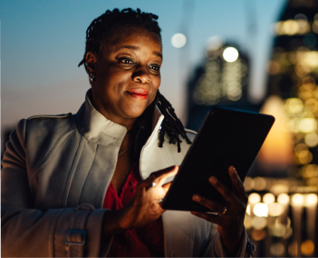 Person holding a tablet outdoors at night, city lights blurred in background, wearing a coat and standing near illuminated buildings