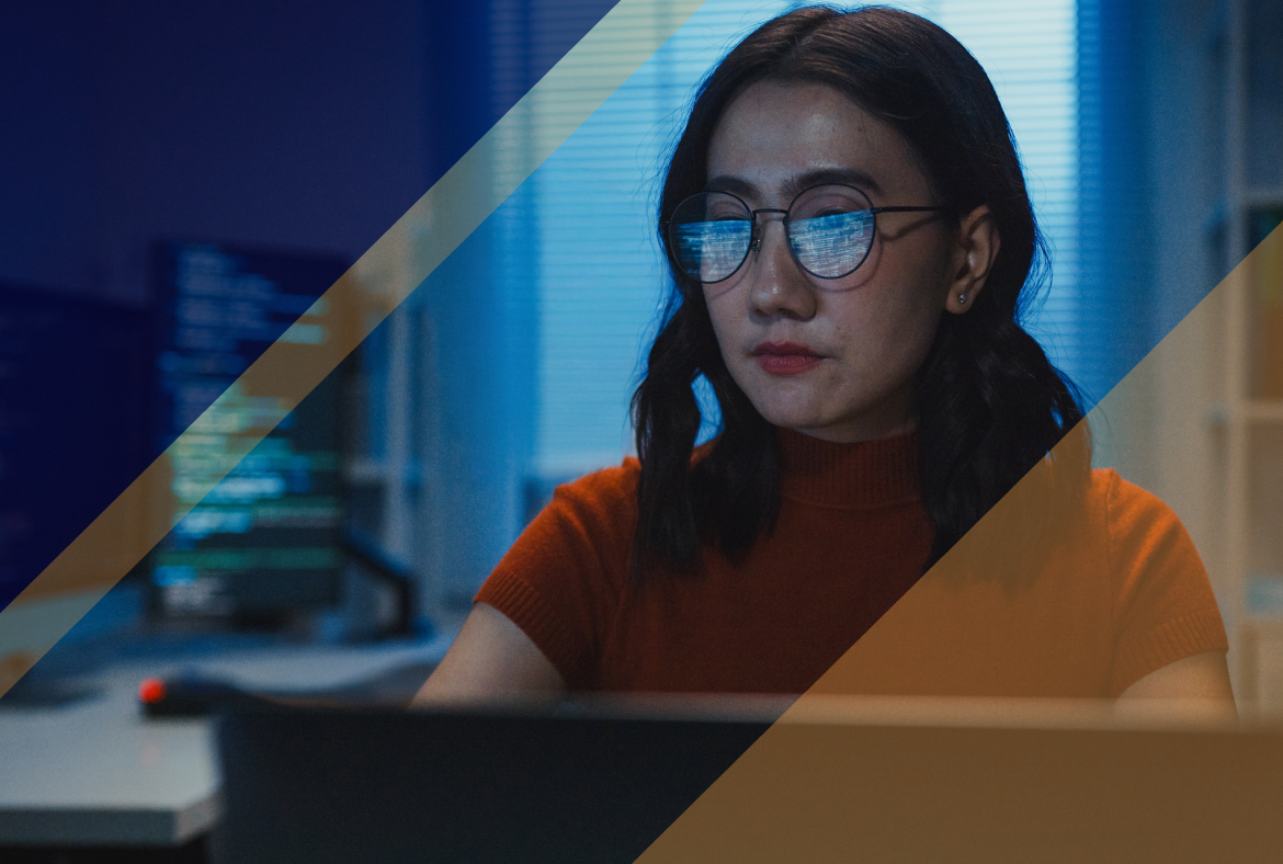 Person wearing glasses working at a laptop, with code reflected on the screen, in a dimly lit office suggesting active security monitoring
