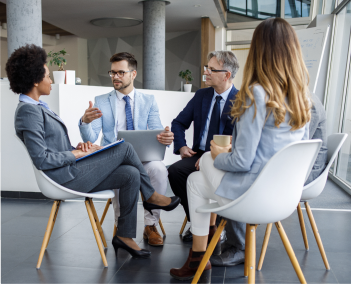 Four professionals seated in a modern office meeting area, discussing documents and laptops in a collaborative business meeting.