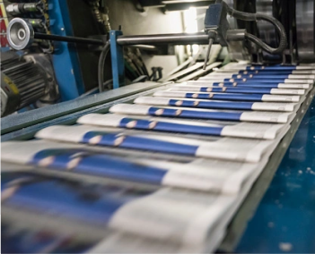 Newspapers moving along a printing press conveyor in a production facility, with stacks of freshly printed papers in motion.