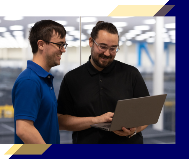 Two people standing indoors review a laptop together in a bright data center or technical workspace, with server racks and overhead lighting in the background.