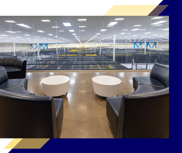 Seating area with black chairs and round white tables overlooking a large data center floor filled with server racks and yellow cable trays behind glass.