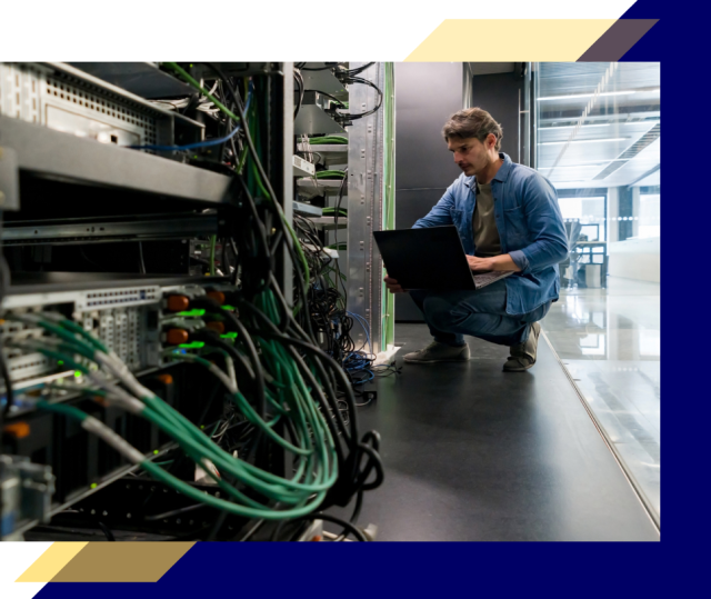 Person kneels beside a server rack using a laptop, surrounded by cables and networking equipment inside a modern data center workspace.