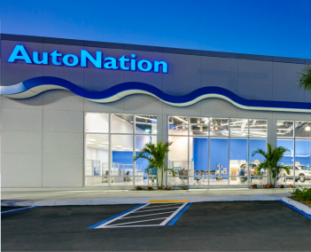 AutoNation car dealership exterior at dusk, with illuminated logo, large glass showroom windows, parked cars, and palm trees in front