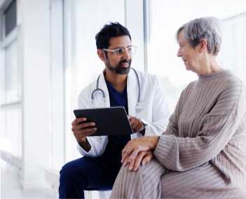 Person in white coat with stethoscope reviews a tablet with seated person in knit outfit near a bright window.