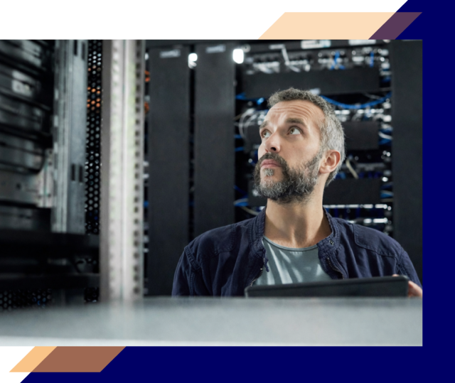 Person holding a tablet while looking up at server racks in a data center, surrounded by network equipment and cabling
