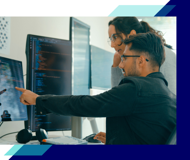 Two people collaborate at a desk, one pointing at multiple monitors displaying code while the other observes, working together at a computer workstation.