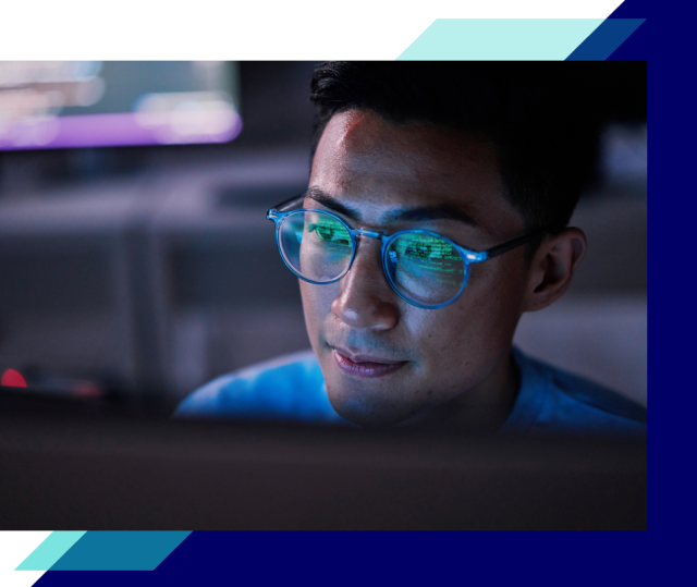 Close-up of a person wearing blue glasses, code reflected in the lenses, looking at a computer screen in a low-lit workspace
