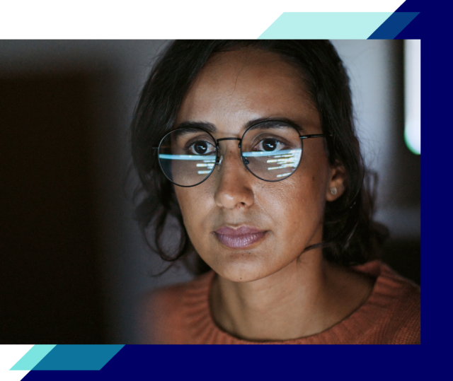 Close-up of a person wearing glasses, with digital code reflected in the lenses, focused on a screen in a dimly lit workspace
