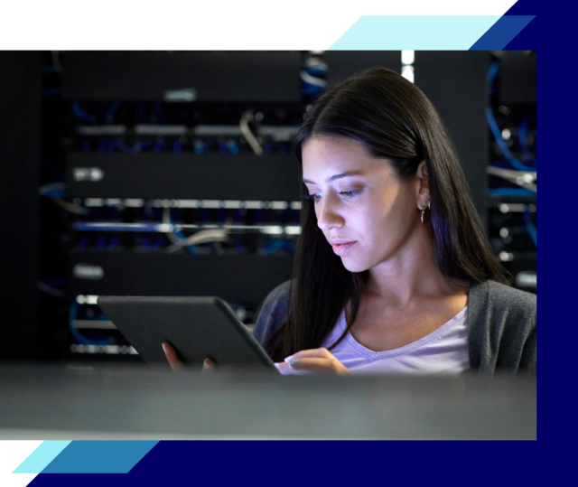 Person holding a tablet in a data center, standing in front of server racks with cables and equipment visible in the background