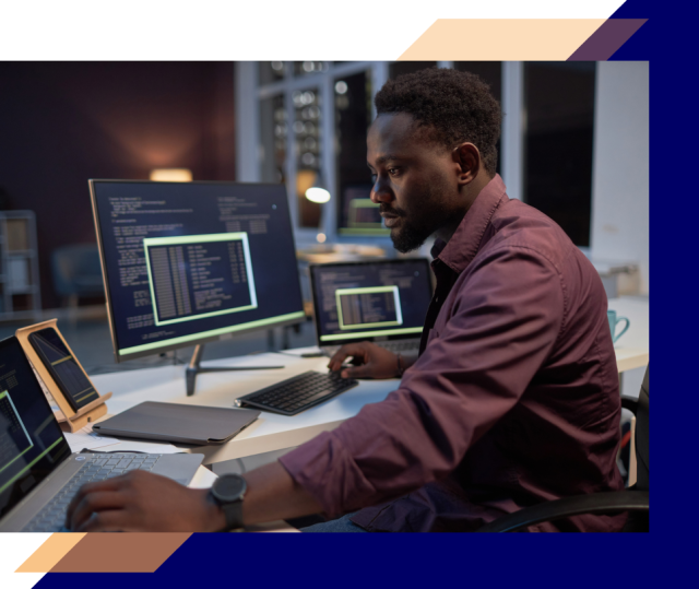 Person working at a desk with a laptop and multiple monitors displaying code and security dashboards in a modern office environment