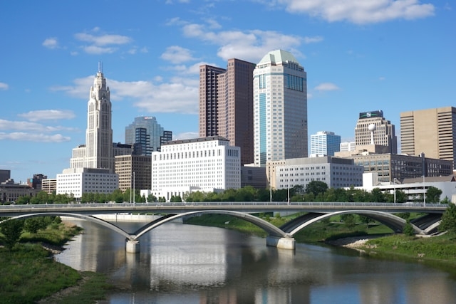 Daytime Columbus, OH skyline with modern and historic buildings beyond an arched bridge spanning a calm river under a blue sky.