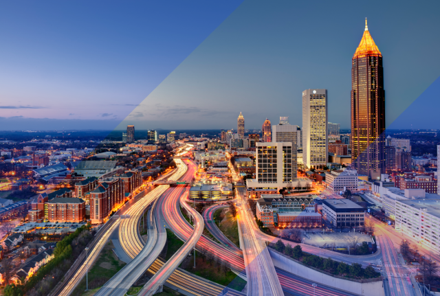 Aerial view of a city skyline at dusk with illuminated highways, light trails from traffic, and tall buildings against a fading blue sky