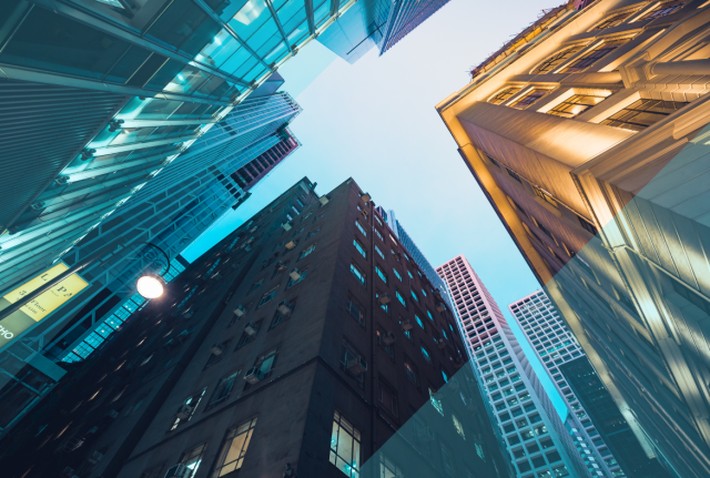 Upward view between tall city buildings with glass and stone facades, showing contrasting blue and warm tones against a bright sky