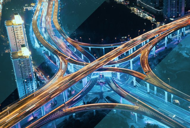 Aerial view of a brightly lit highway interchange at night, with intersecting roads and light trails from traffic amid city buildings