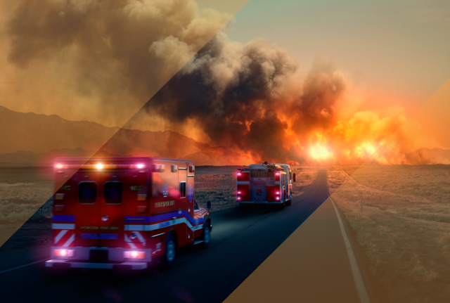 Two fire trucks drive along a road toward a large wildfire, with flames and thick smoke rising across a dry landscape under an orange sky