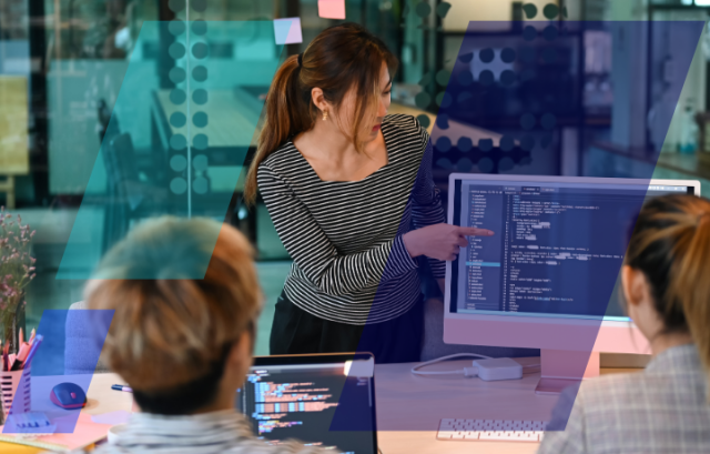 Person presenting code on a desktop screen to a small group in a modern office, with laptops and colorful glass panels in the background