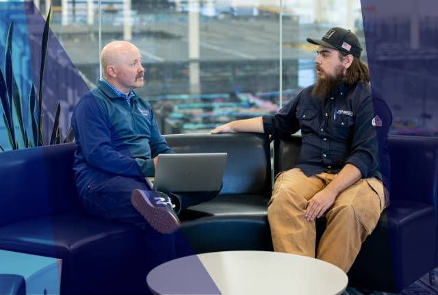Two individuals seated on a couch in an office lounge, conversing beside a laptop, with a glass wall and data center visible behind them
