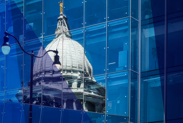 Reflection of a domed government building on a blue glass facade, with a streetlamp visible in the foreground