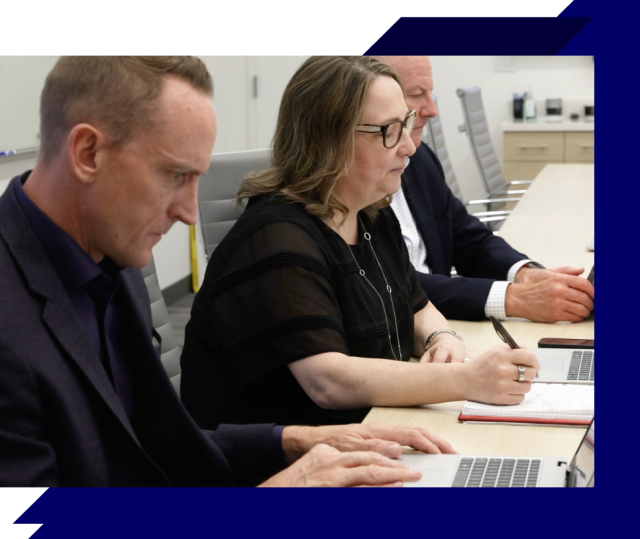 Three people at a conference table using laptops and taking notes in a meeting room with a neutral office setting and blue graphic accents