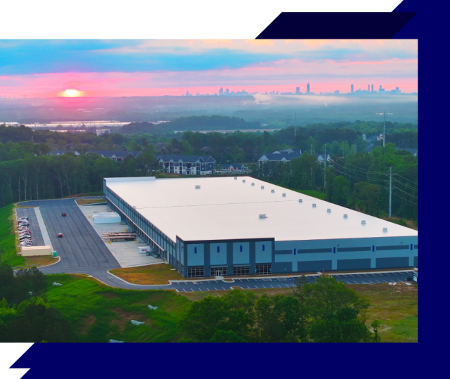 Aerial view of a large warehouse building at sunrise with surrounding greenery and a distant city skyline on the horizon