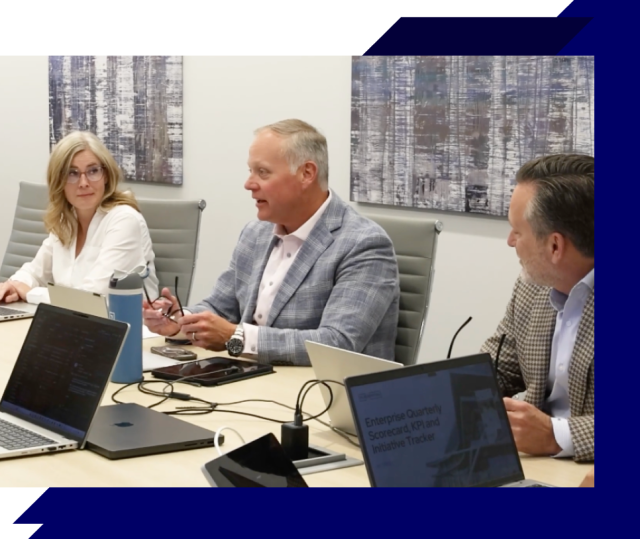Three people in a conference room meeting, seated at a table with laptops and discussing, with abstract artwork on the wall behind