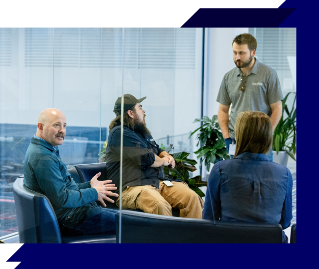 Four people in a glass-walled office, seated and standing around a sofa, engaged in a casual team discussion.