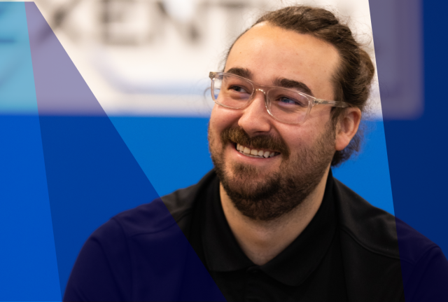 Close-up of person wearing glasses, seated against a blue background with abstract overlay, looking to the side and smiling gently