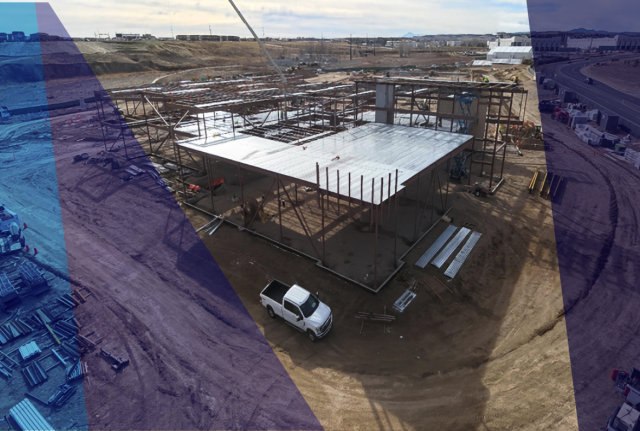 Aerial view of a construction site with steel framing, concrete foundation, and a white work truck on a dirt lot surrounded by open land