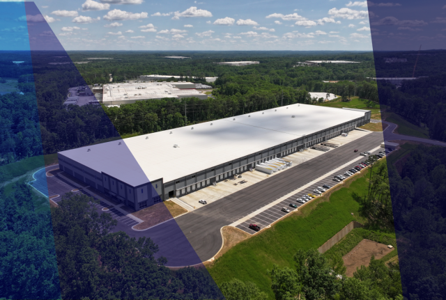Aerial view of a large industrial building with parking lots, loading docks, and surrounding forest under a partly cloudy sky