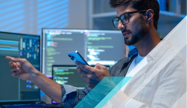 Person using a smartphone at a workstation with multiple code-filled monitors, featuring blue geometric overlays.