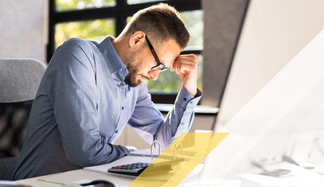 Person seated at a desk, holding their forehead while looking down beside a computer monitor, with geometric white and gold graphic overlays.