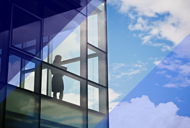 Person standing inside a glass office building, viewed through large windows against a bright blue sky with geometric blue overlays.