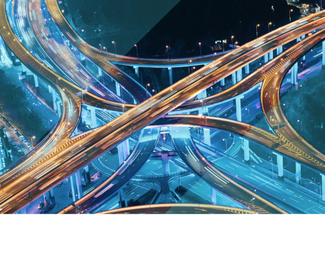Aerial view of a brightly lit highway interchange at night, with glowing traffic trails and blue geometric overlays symbolizing IT agility.