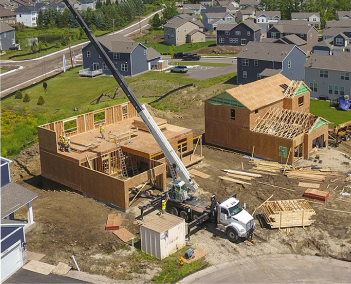 Aerial view of two houses under construction with a crane, framing, and building materials in a suburban neighborhood